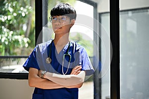 A positive Asian male doctor stands in the hospital corridor with his arms crossed