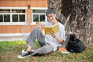 A positive Asian male student is sipping coffee and reading a book under the tree in a campus park