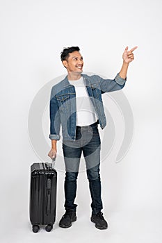 A positive Asian man with his luggage is pointing his finger at an empty space, isolated background