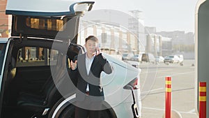 Positive asian man in stylish business wear talking on mobile phone while standing near electric car at outdoors