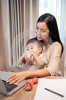 A positive Asian mom is feeding milk from a bottle to her baby boy while working on her laptop