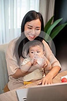 A positive Asian mom is feeding milk from a bottle to her baby boy while working on her laptop