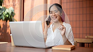 A positive Asian woman is talking on the phone with her client while working remotely at a cafe