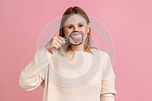 Positive attractive blond woman standing, holding magnifying glass on his teeth, looking at camera.