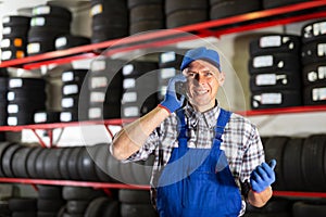 Positive auto mechanic talking on a mobile phone against the background of car tires in car service