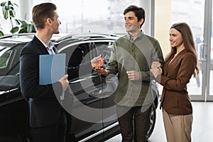 Positive auto showroom manager giving car key to happy young clients at car dealership