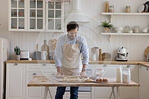 Positive baker man wearing apron rolling dough on floury table