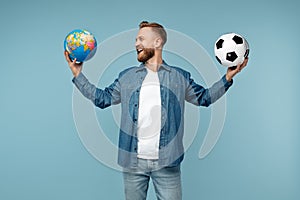 Positive bearded blonde hipster man in medical mask holding classic soccer ball and world globe over blue background.