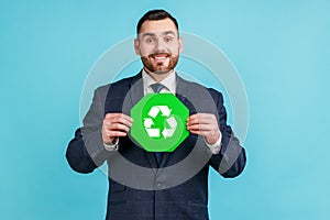 Positive bearded man wearing official style suit holding green recycling sign in hand and looking