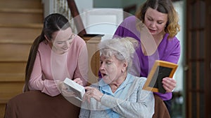 Positive beautiful Caucasian senior woman telling stories to daughter and granddaughter looking at pictures. Portrait of