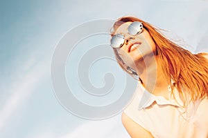 Positive beautiful happy red haired girl in the mirror sunglasses on blue sky background, summer sunset time