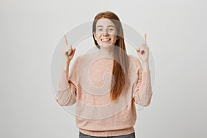 Positive beautiful redhead european woman pointing up with index fingers while smiling broadly and glancing at camera