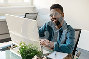 Positive black businessman in eyeglasses smiling at camera, sitting in front of computer at workdesk in office