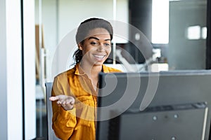 Positive black female entrepreneur gesticulating at computer webcamera, working and having video call in office