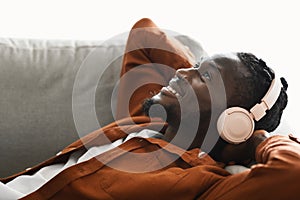 Positive black man in headphones lying on sofa and listening to music, dreaming and relaxing with hands behind head