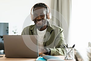 Positive black man in wireless headphones working online on laptop at home office, looking and smiling at camera