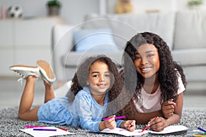 Positive black mom and her happy daughter drawing together on floor at home. Parent child pastimes concept