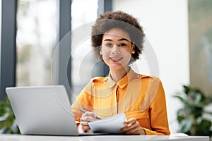 Positive black teenage student girl watching online webinar on laptop and writing notes, studying in university library