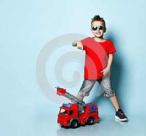 Positive blond boy in bright stylish casual clothing, sneakers, sunglasses standing near toy fire engine and showing thumb sign