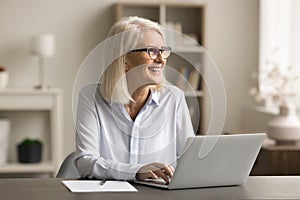 Positive blonde elder freelancer woman in eyeglasses working at home