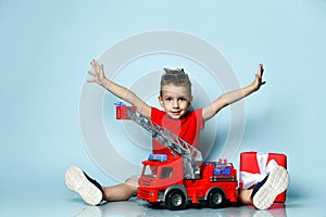 Positive boy in bright stylish casual clothing and sneakers sitting on floor with toy fire engine and feeling happy