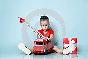 Positive boy in bright stylish casual clothing and sneakers sitting on floor with toy fire engine and feeling happy