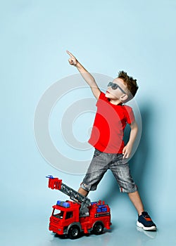 Positive boy in bright stylish casual clothing, sneakers, sunglasses standing near toy fire engine and pointing up with finger