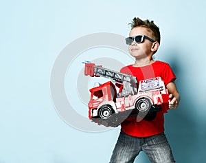 Positive boy in bright stylish casual clothing and sunglasses standing and holding toy fire engine in hands over blue background