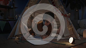 Positive boy drawing with colored pencils on white paper shit while lying on floor in decorative tent at home. Male teen