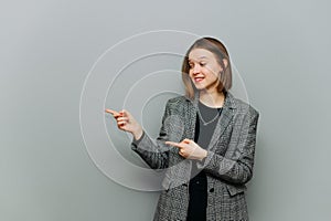 Positive business lady in a jacket stands on a gray background with a smile on his face looks and points his finger to the side in