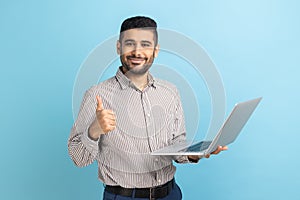 Positive businessman standing with portable computer in hand, looking at display, showing thumb up.