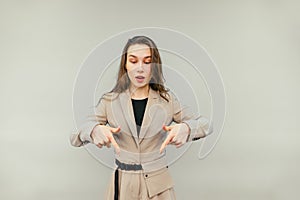 Positive businesswoman in a suit stands on a beige background and shows fingers down on an empty spot with a surprised face