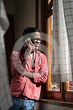 Positive carefree African American man student standing in room next to window and talking on phone