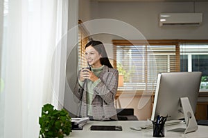 Positive caucasian businesswoman holding cup of coffee and looking away standing at home office