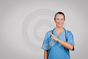 Positive caucasian female doctor surgeon in blue uniform pointing finger at copy space on gray background