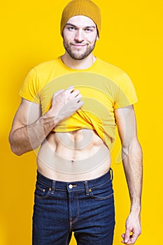 Positive Caucasian Handsome Man In Yellow Tshirt Posing in Warm Hat Over Yellow Background While Pulling Shirt Off