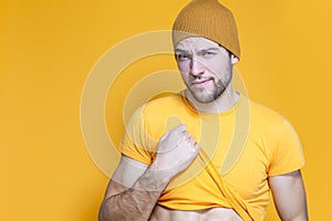 Positive Caucasian Handsome Man In Yellow Tshirt Posing in Warm Hat Over Yellow Background While Pulling Shirt Off