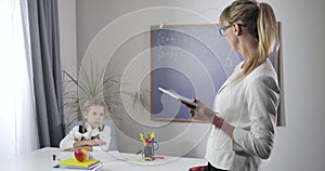 Positive Caucasian schoolgirl making victory gesture as sitting at the table. Adult female tutor teaching girl at home