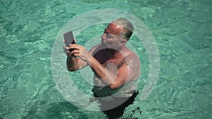 Positive Caucasian tourist gesturing thumb up in slow motion standing in turquoise sea talking on phone. Portrait of