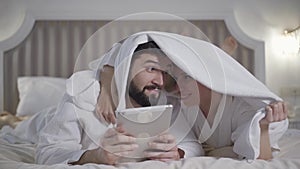 Positive Caucasian woman distracting husband or boyfriend from tablet covering him with towel. Happy loving couple