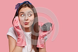 Positive Caucasian woman pretends speaking on mobile phone, holds sponge near ear, dressed in casual clothes, wears