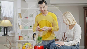 Positive Caucasian woman sitting on table as Middle Eastern man cooking healthful smoothie in blender. Cheerful