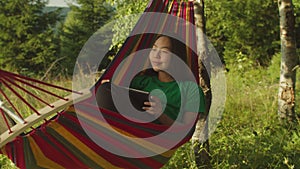 Positive charming asian female traveler resting in hammock, working with digital tablet in nature