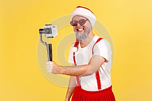 Positive cheerful man in santa claus costume and stylish sunglasses posing on camera of smartphone in his hand, blogger recording