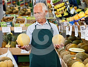 Positive senior male owner of greengrocery shop in apron offering fresh fruits and vegetables