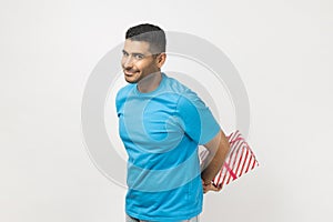 Positive cheerful unshaven man with present box behind back, gift for holiday, congratulating.