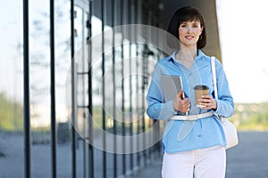 Positive confident businesswoman in blue shirt holds digital tablet and coffee drink in paper cup outside of modern