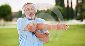 Positive confident senior sportsman wearing wireless headphones doing arm stretching in park outside