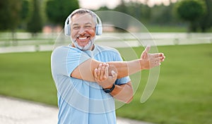 Positive confident senior sportsman wearing wireless headphones doing arm stretching in park outside