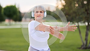 Positive confident senior sportswoman wearing wireless headphones doing arm stretching in park outside